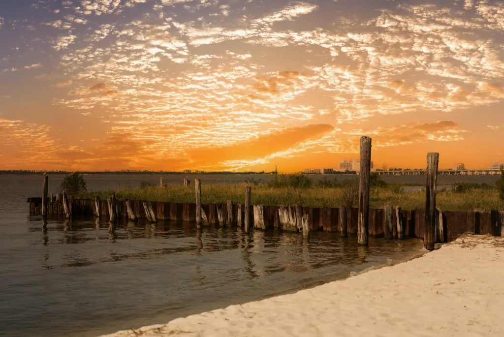 Sunset over a Gulf Coast shoreline with a wooden bulkhead, white sand, and the Biloxi skyline in the distance